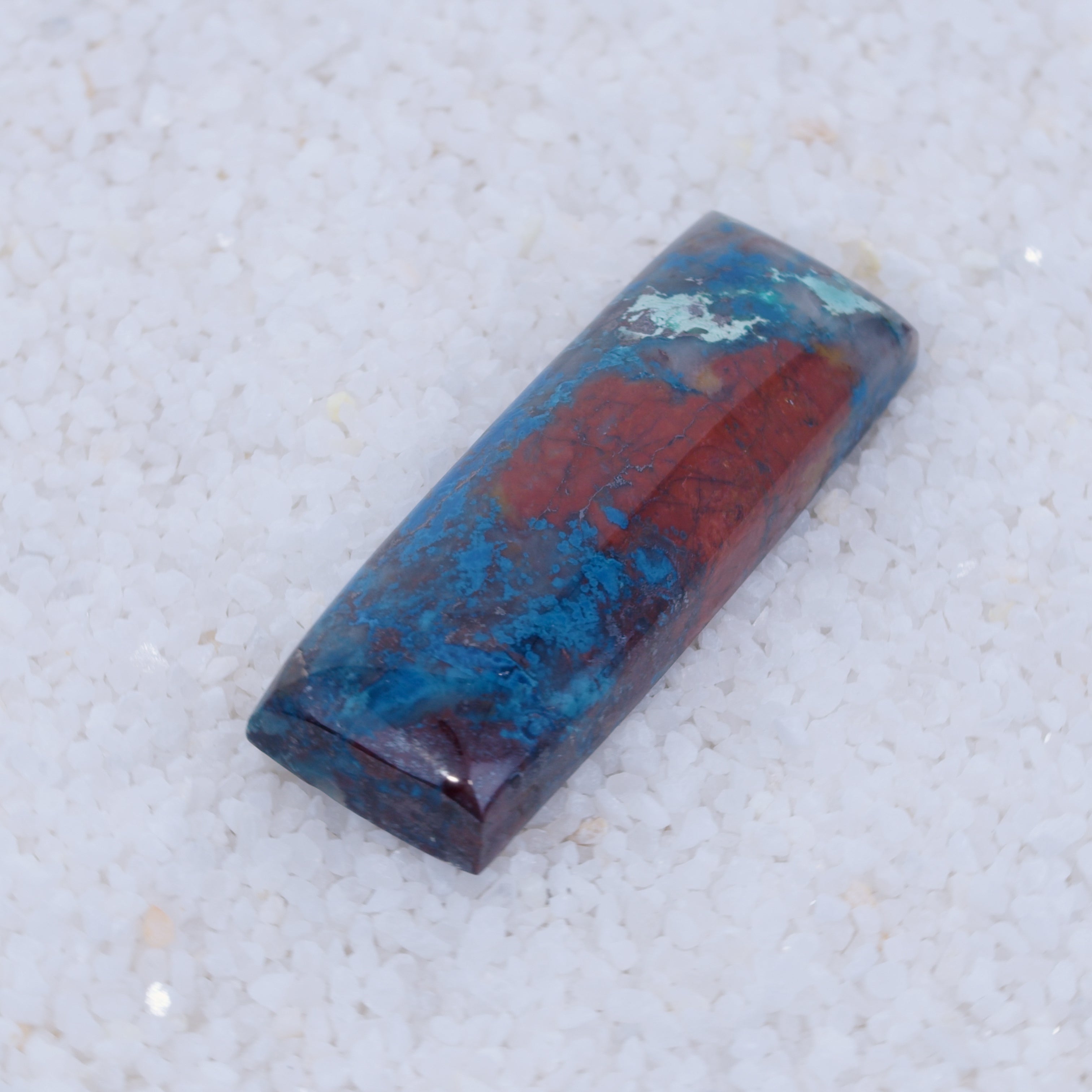 Rectangular Canyonite gemstone with blue and red hues on a light gray background
