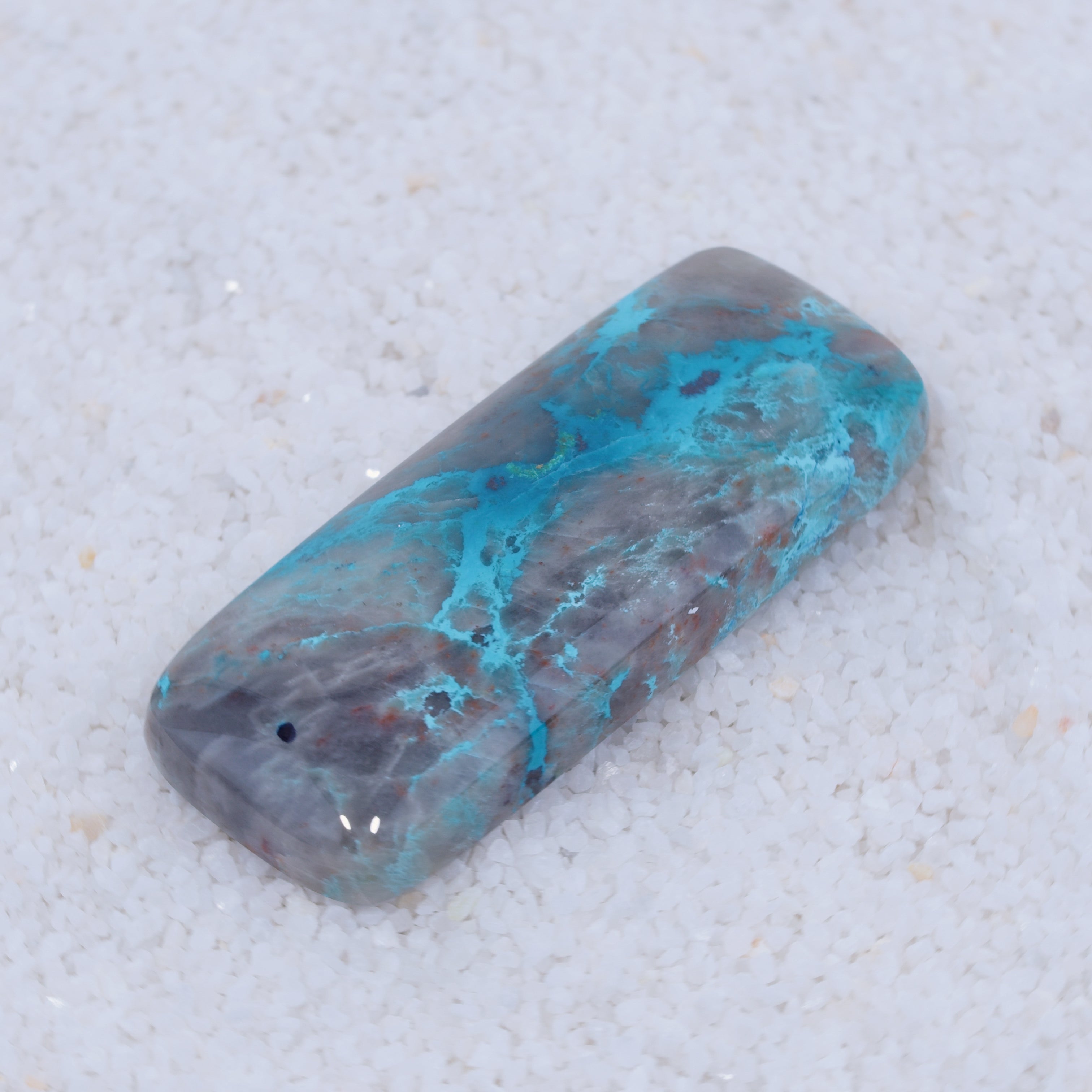 A rectangular Canyonite Sky LTD gemstone with a marbled mix of gray and vibrant turquoise blue streaks rests on fine white sand. It features rounded edges, a glossy reflective surface, and is ideal for collectors.