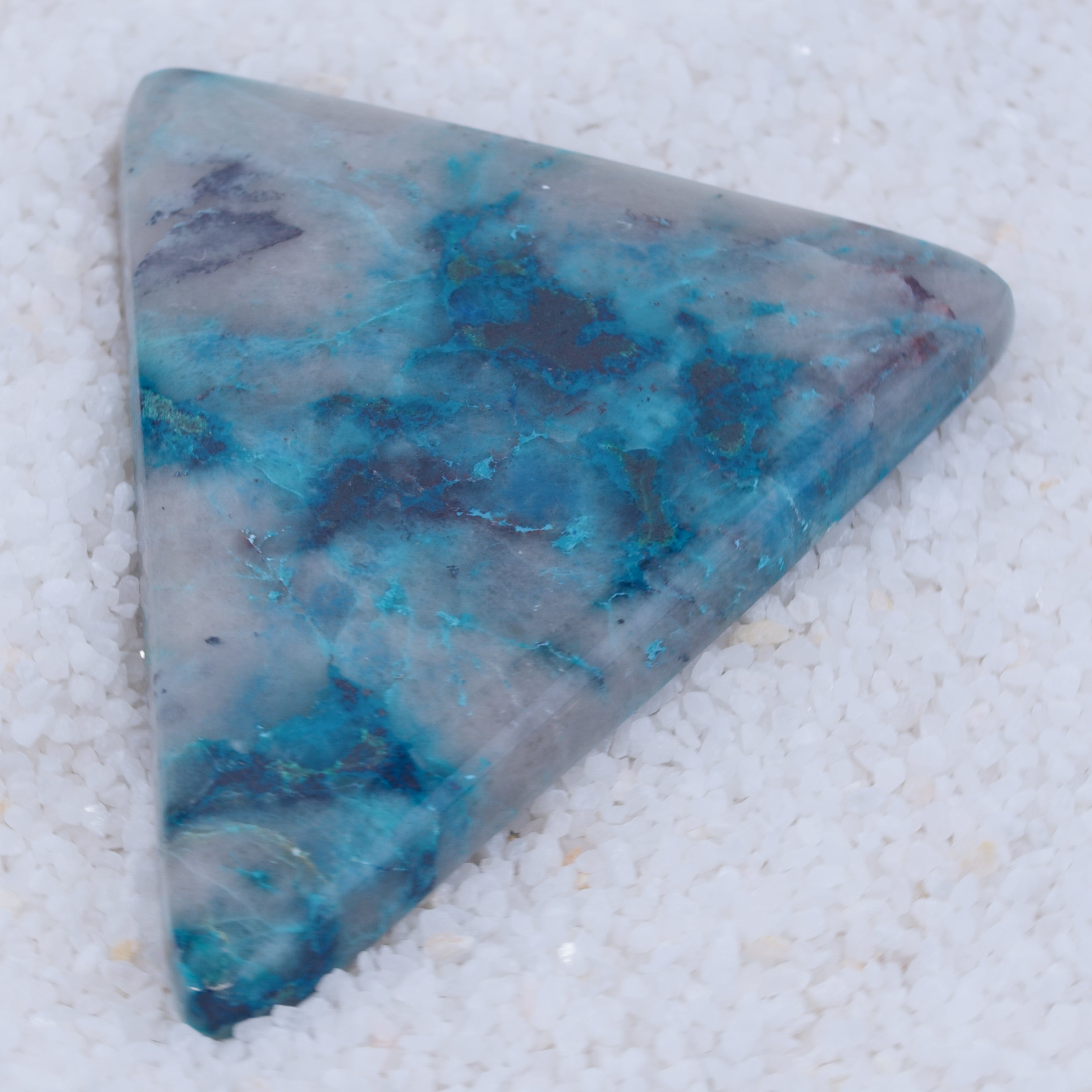 A polished, triangular Canyonite Sky LTD gemstone with a marbled blue, teal, and gray pattern rests on white crystals. Its smooth surface highlights vibrant hues, making it a rare collector’s piece.