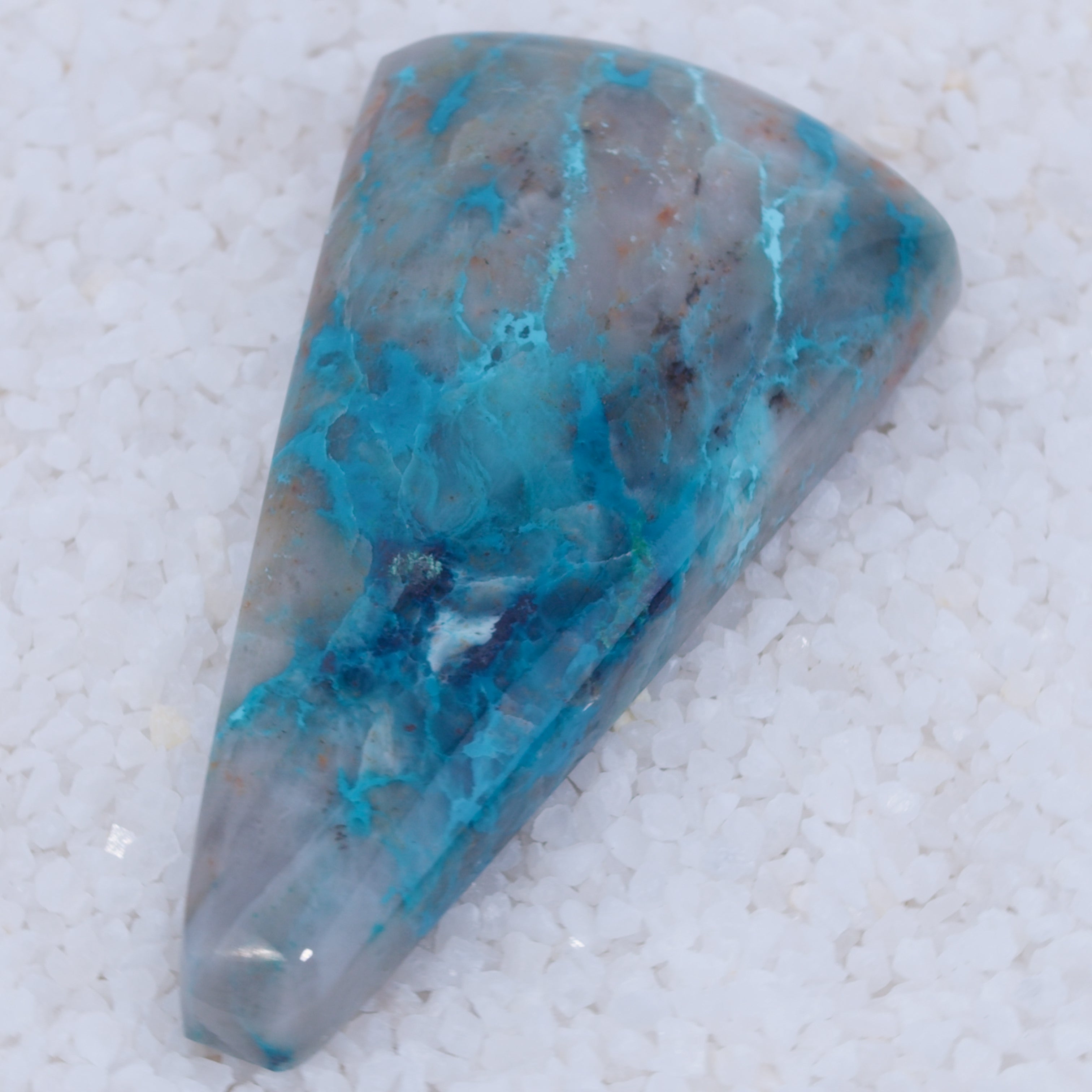 A rare collector’s gemstone from Canyonite Sky LTD, this polished triangular Canyonite Sky gem displays swirling blue, turquoise, gray, and hints of brown on a smooth surface. Vibrant colors stand out as it rests on white pebbles.