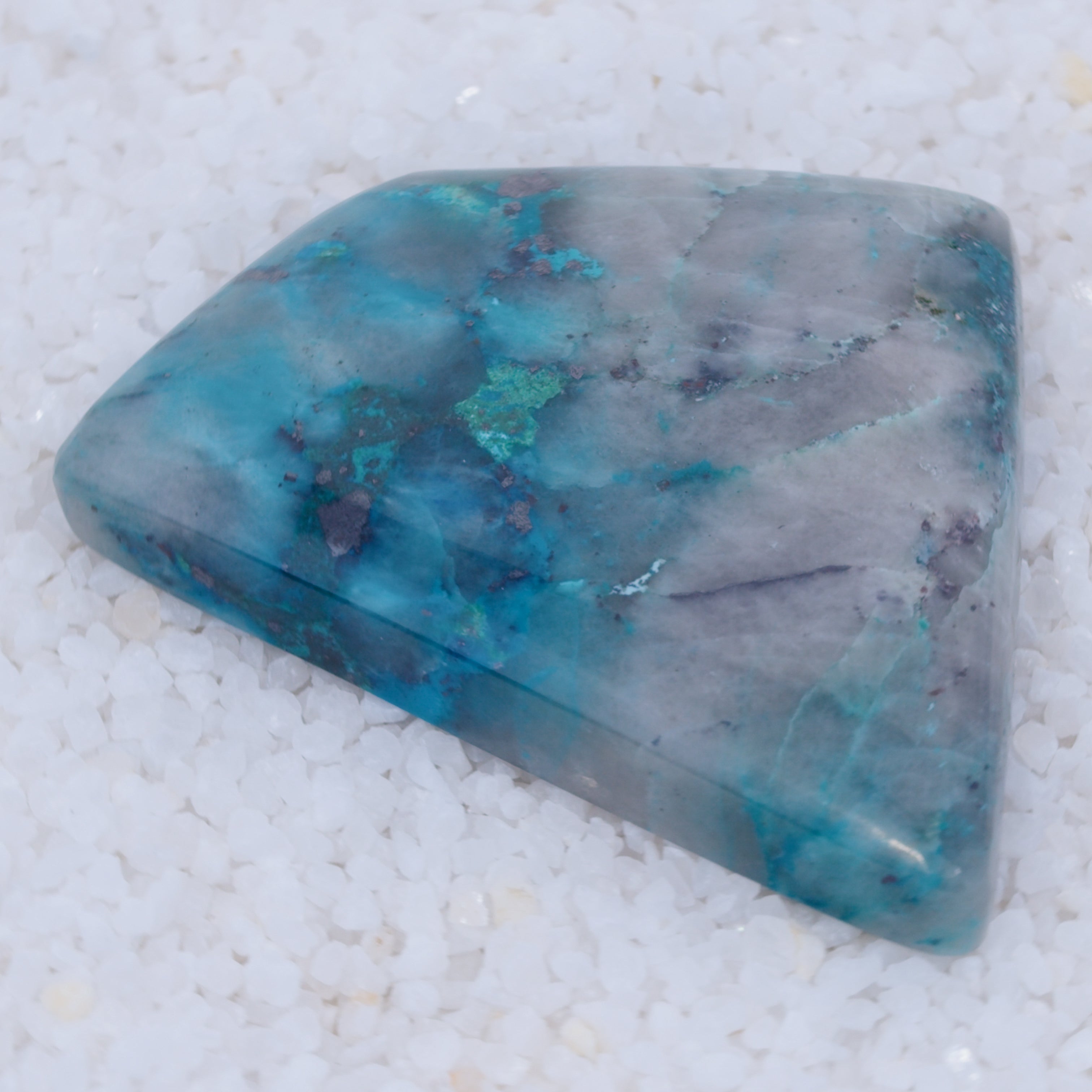 A polished, triangular Canyonite Sky LTD cabochon displays blue, teal, and white marbled patterns. This rare collector’s gem rests on fine white crystals, highlighting its smooth surface and striking colors.