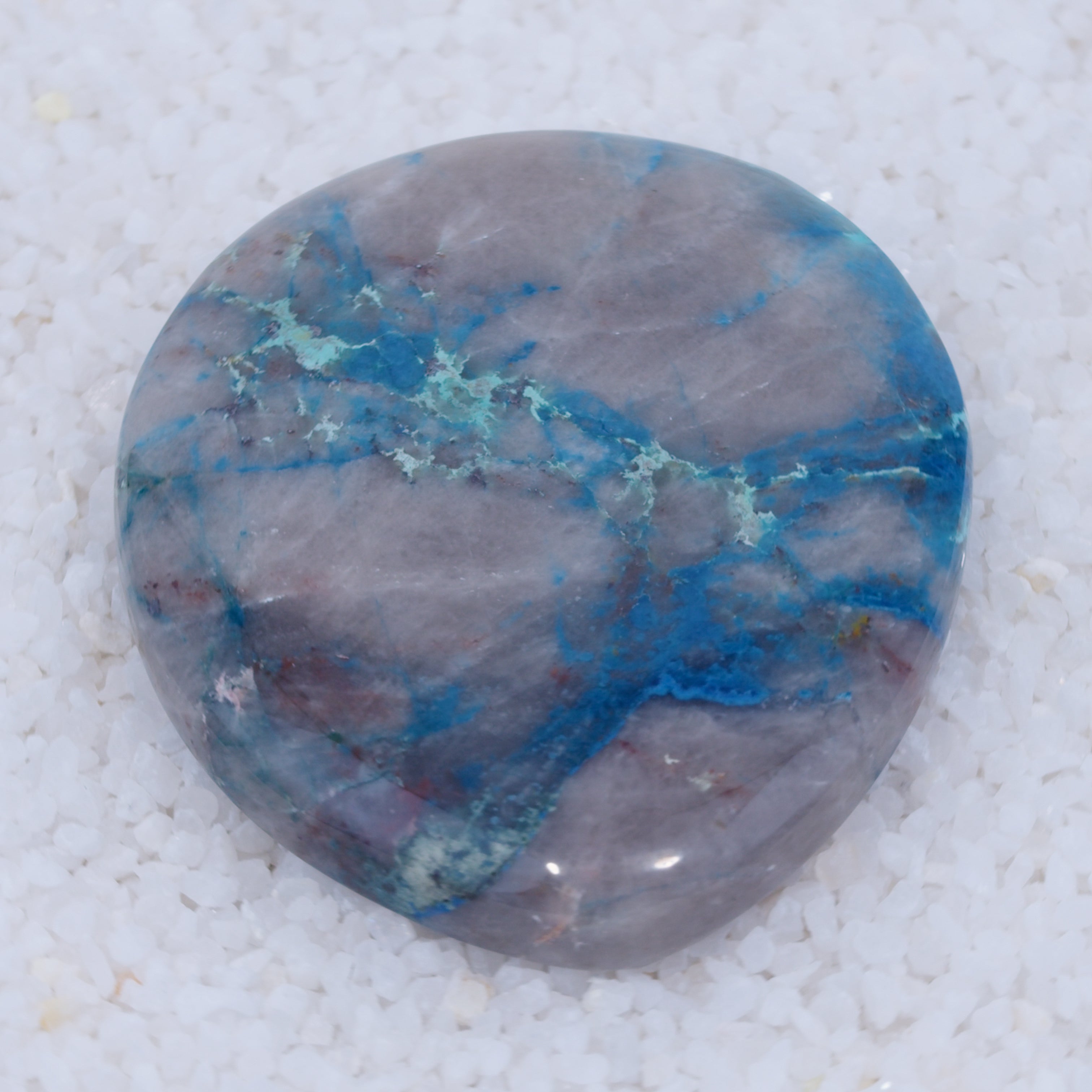 A polished, oval Canyonite Sky LTD™ gemstone with a sleek, glossy surface. Predominantly gray, it displays striking blue and turquoise veins. Resting on white pebbles, this rare copper silicate cabochon captivates with vivid contrasts.