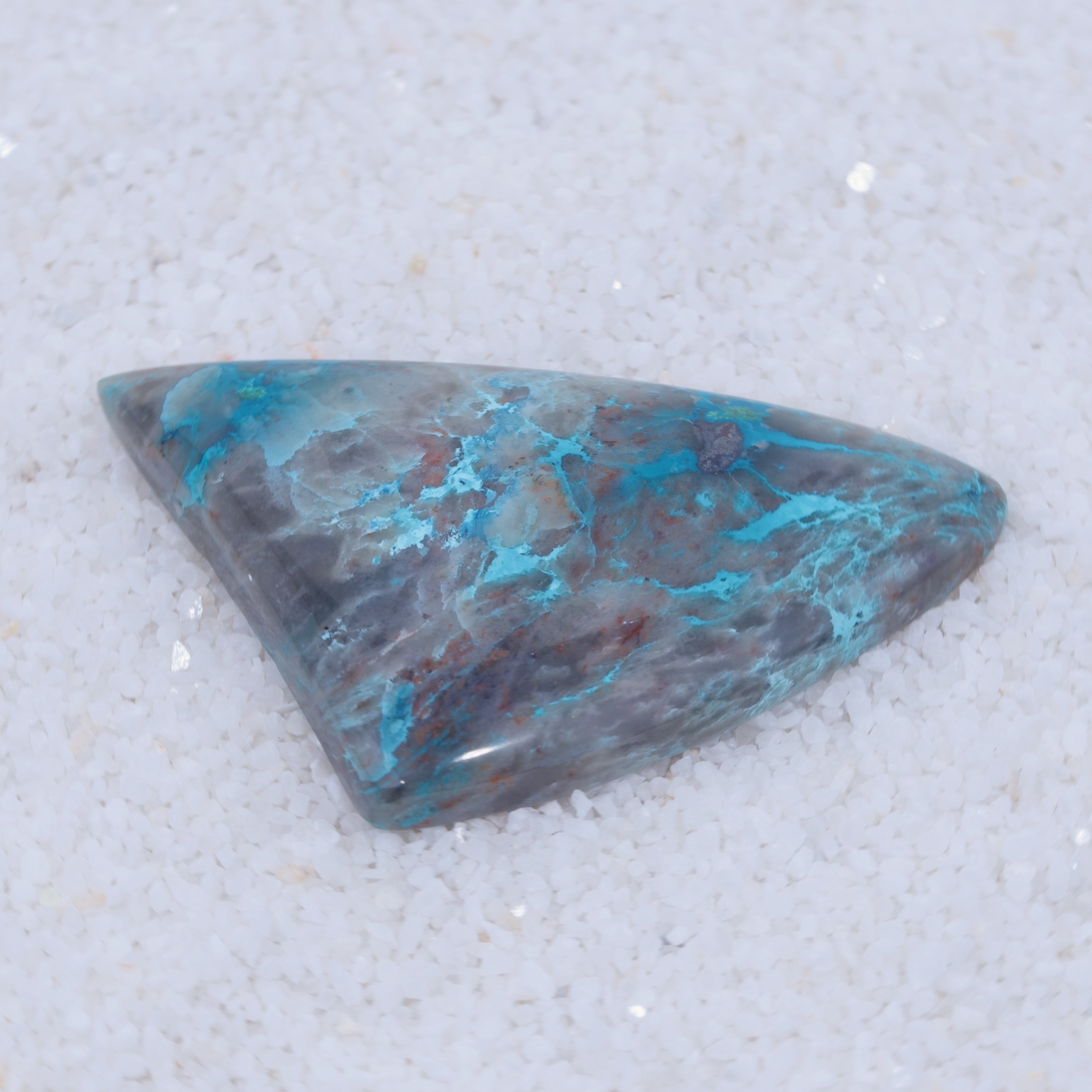 A polished, triangular Canyonite Sky LTD cabochon features blue, teal, grey, and brown marbling on fine white crystals. This rare collector’s gemstone has a glossy finish that accentuates its natural patterns and smooth texture.