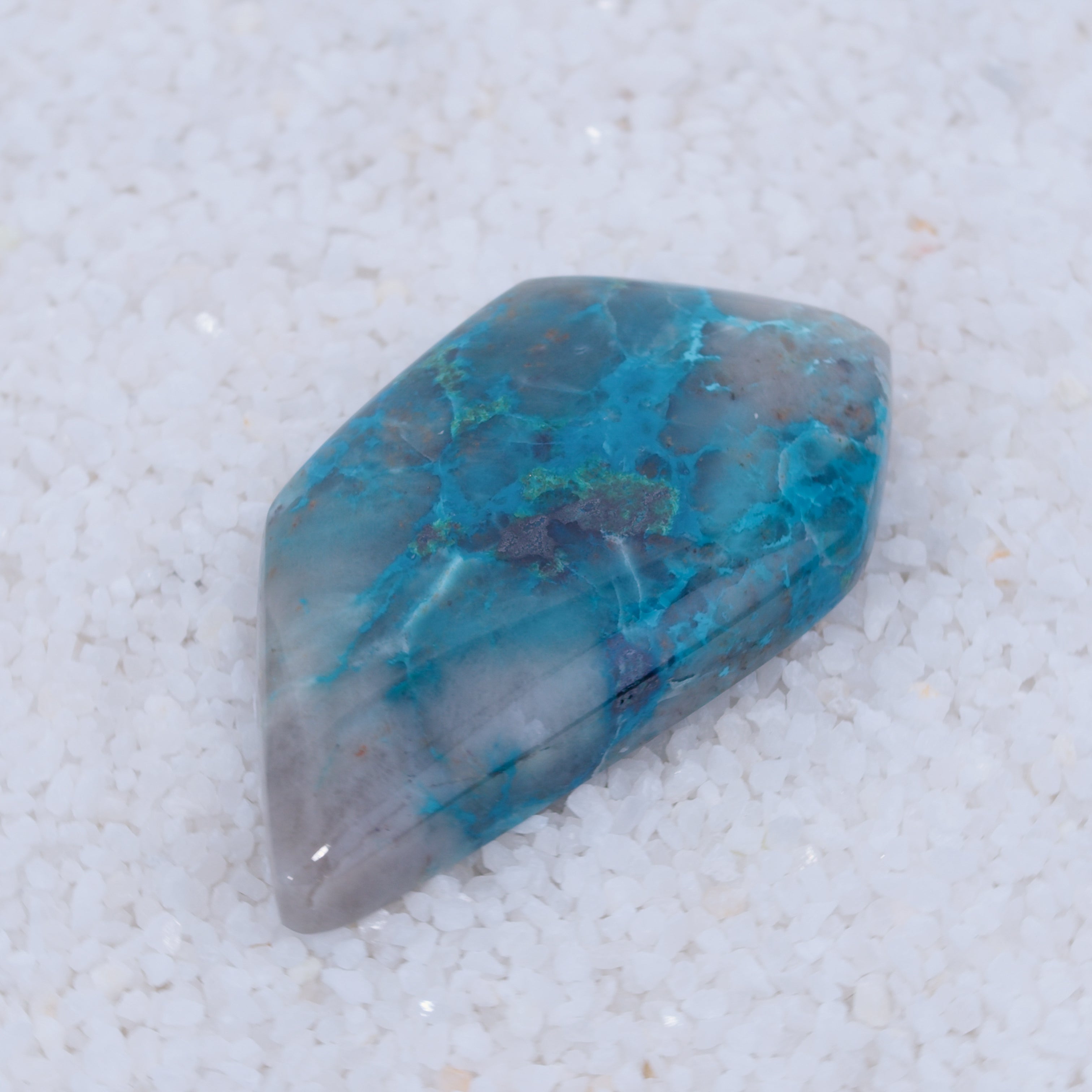A polished, irregular blue cabochon by Canyonite Sky LTD with teal, green, and gray streaks rests on white pebbles. Its smooth, shiny surface shows varied colors and marbling—a unique collector’s gemstone.
