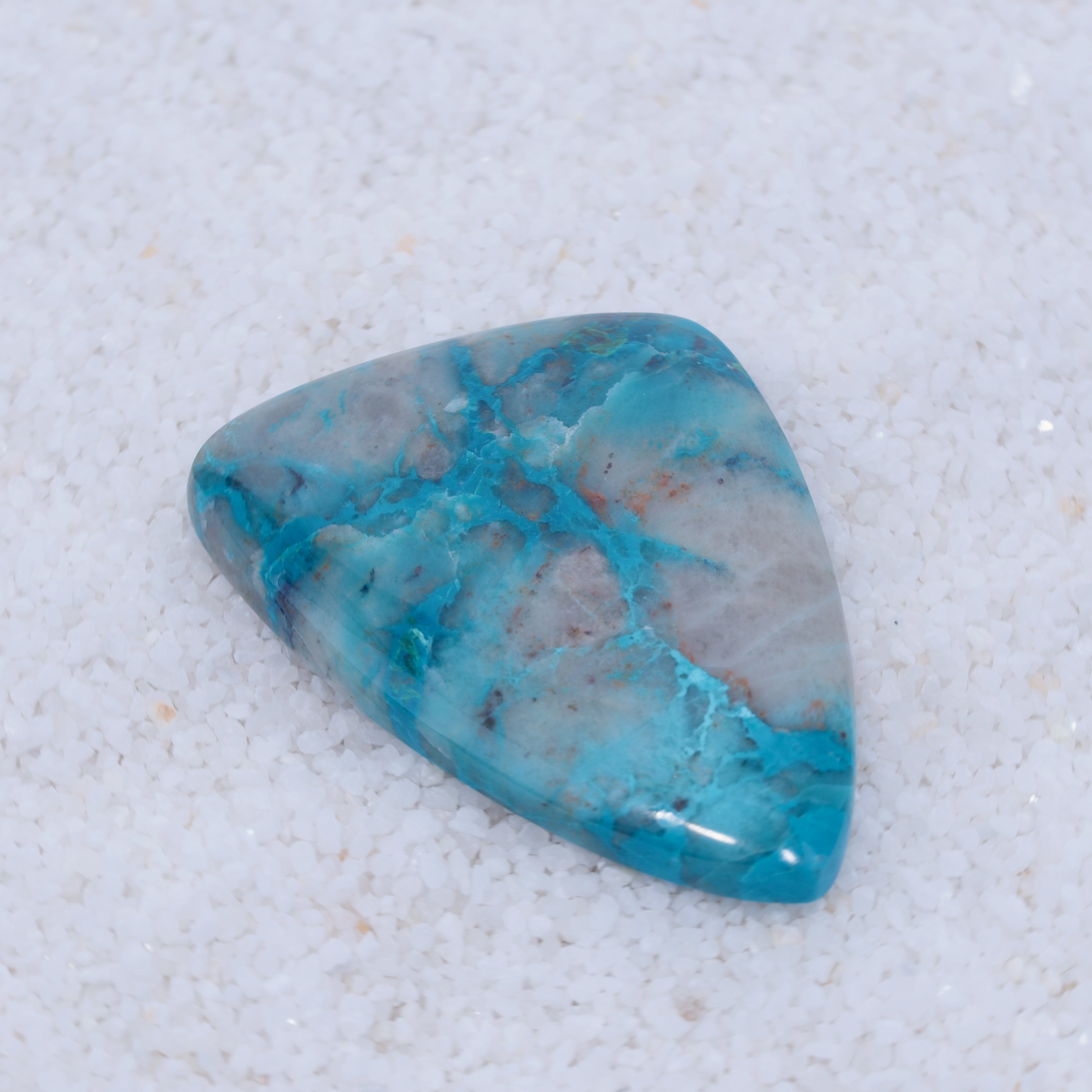 A polished, triangular blue gemstone by Canyonite Sky LTD rests on fine white sand. Its rare cabochon cut features marbled gray and white veins and blue tones from light to deep aqua, making it a collector’s gem.