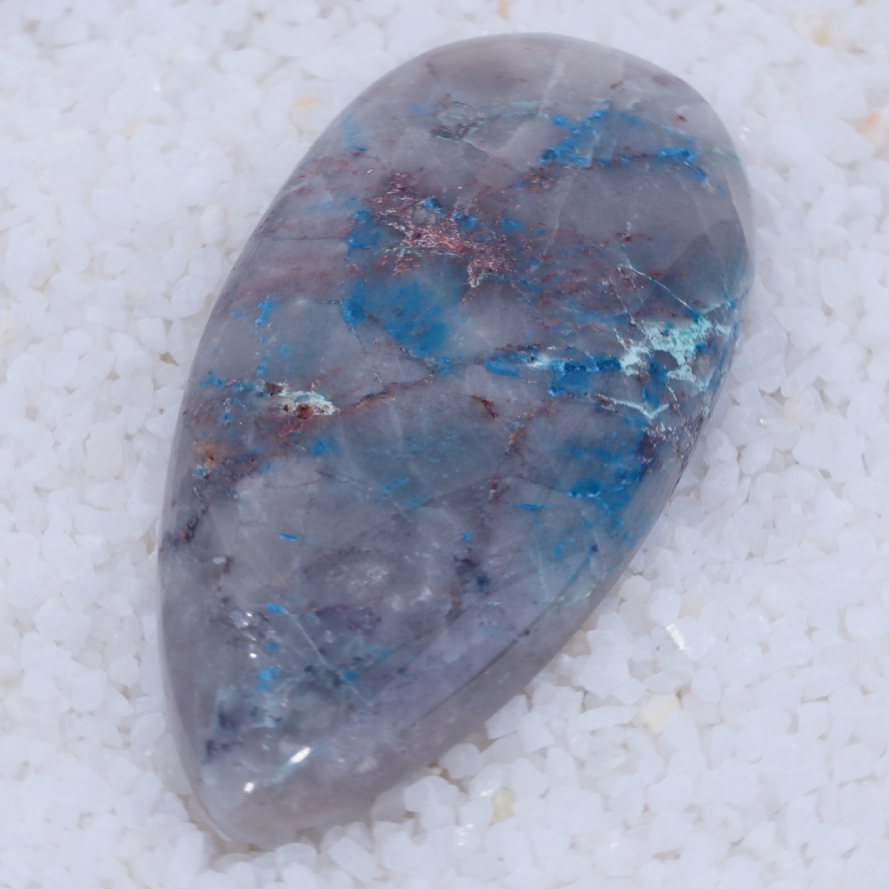 A polished Canyonite Sky cabochon by Canyonite Sky LTD features a gray base, bright blue streaks, and reddish-brown veins. Its smooth surface reflects light atop white pebbles, making it a stunning collector’s gemstone.