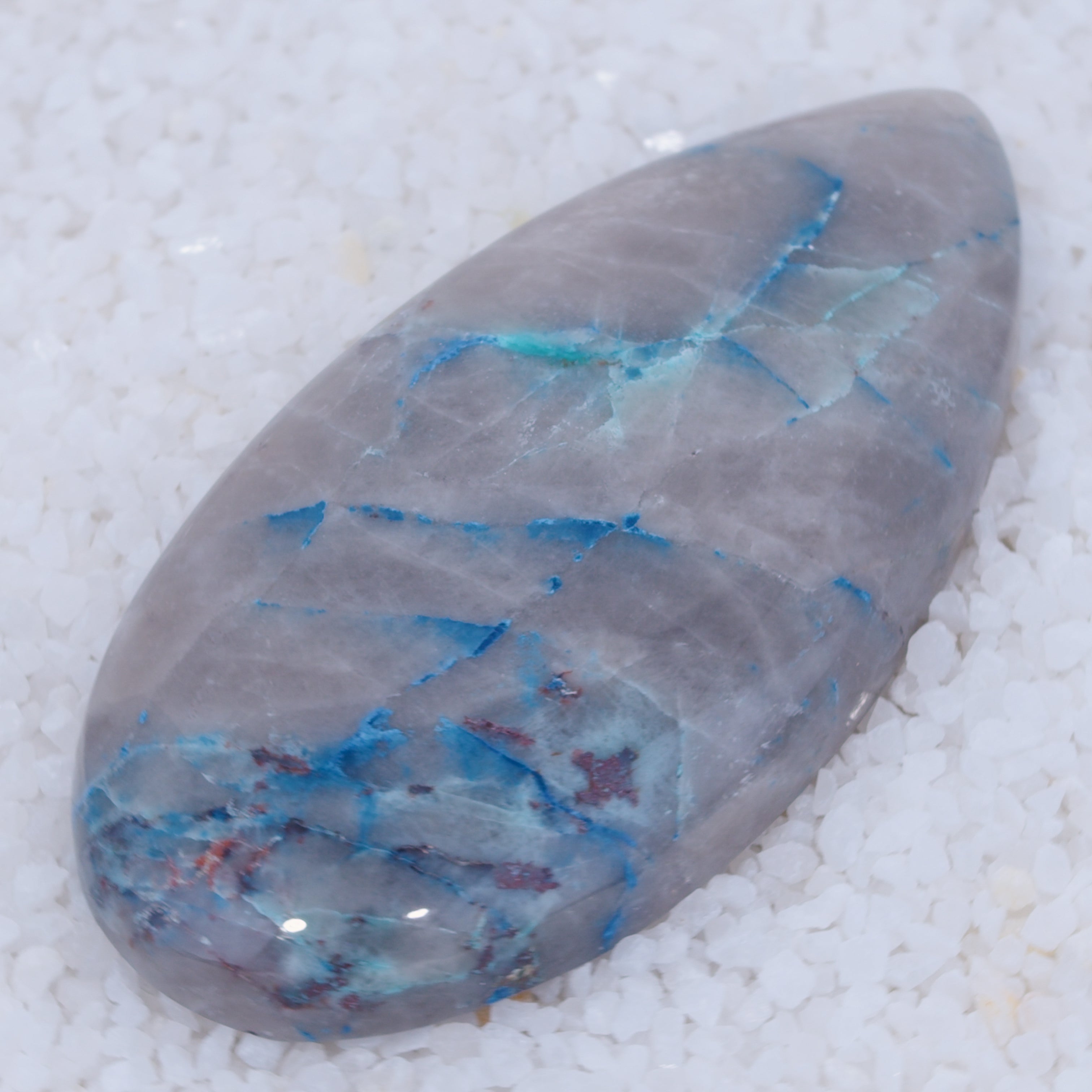 A glossy, oval-shaped gray stone with vivid blue copper silicate veins rests on white crystals. Its natural blue streaks contrast sharply with the gray—a rare cabochon from Canyonite Sky LTD.
