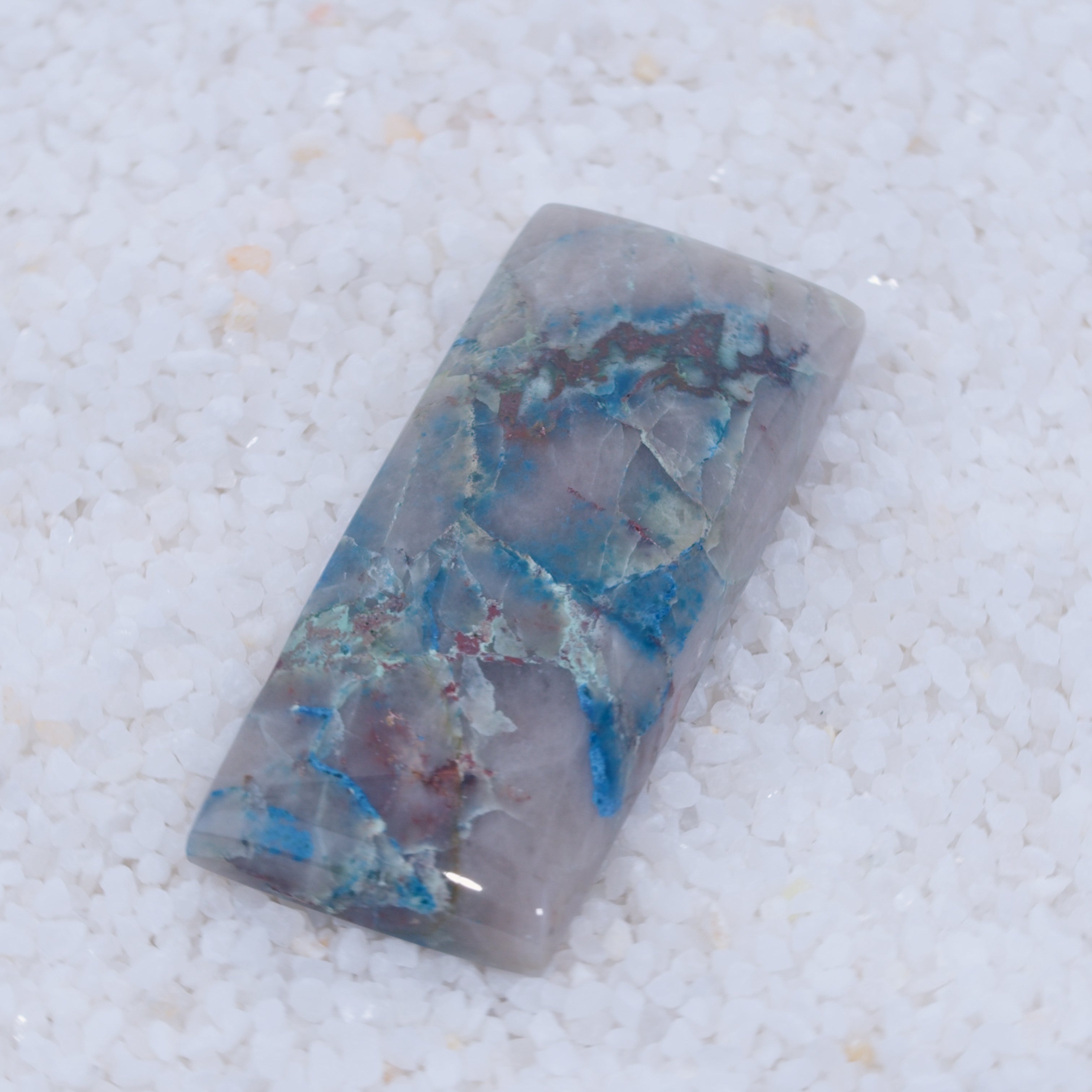 The Canyonite Sky LTD copper silicate cabochon features a rectangular shape with a light gray base, streaks of blue, green, and red, and a glossy surface. Its unique marbled patterns make it a stunning collector’s piece atop white pebbles.