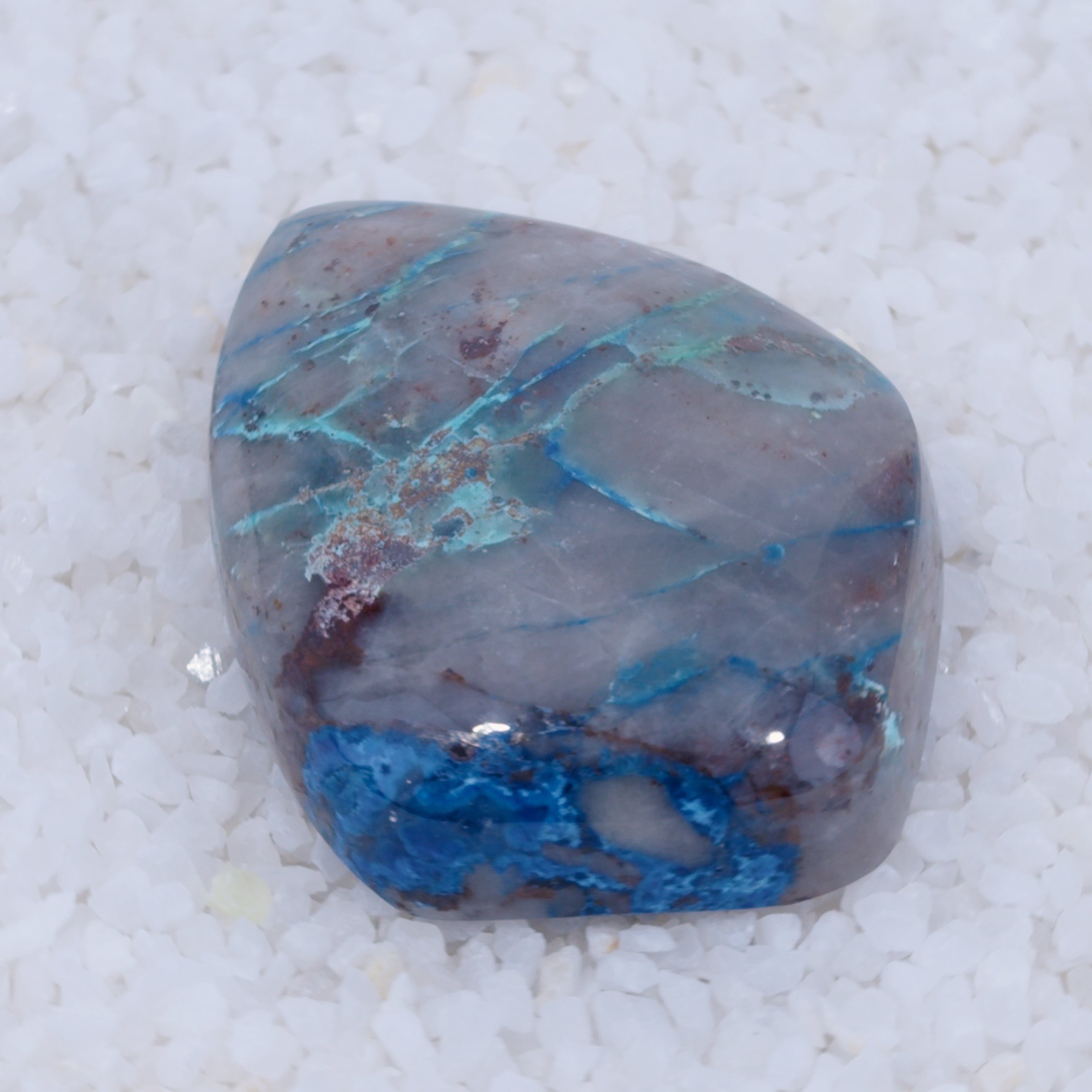 A polished Canyonite Sky LTD cabochon gemstone with a smooth surface rests on white crystals. Its marbled gray base features streaks of bright blue and hints of green and brown from copper silicate minerals for a natural look.
