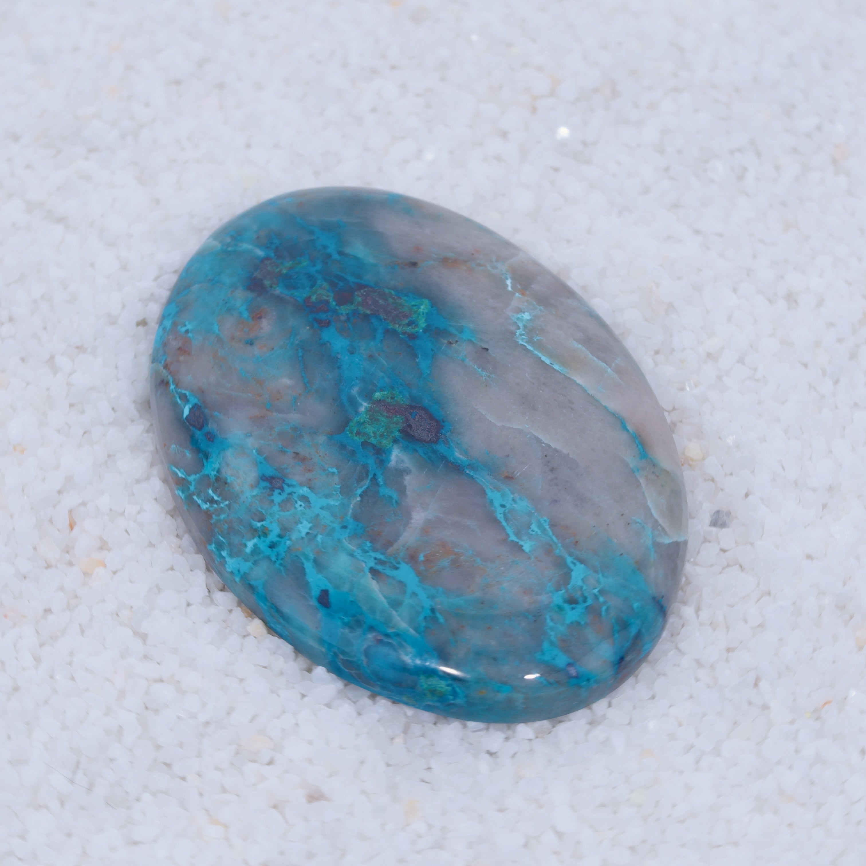 An oval, polished copper silicate cabochon from Canyonite Sky LTD displays swirling blue, teal, and gray patterns with natural veining on a glossy surface—this gemstone is a prized collector’s find resting on fine white sand.