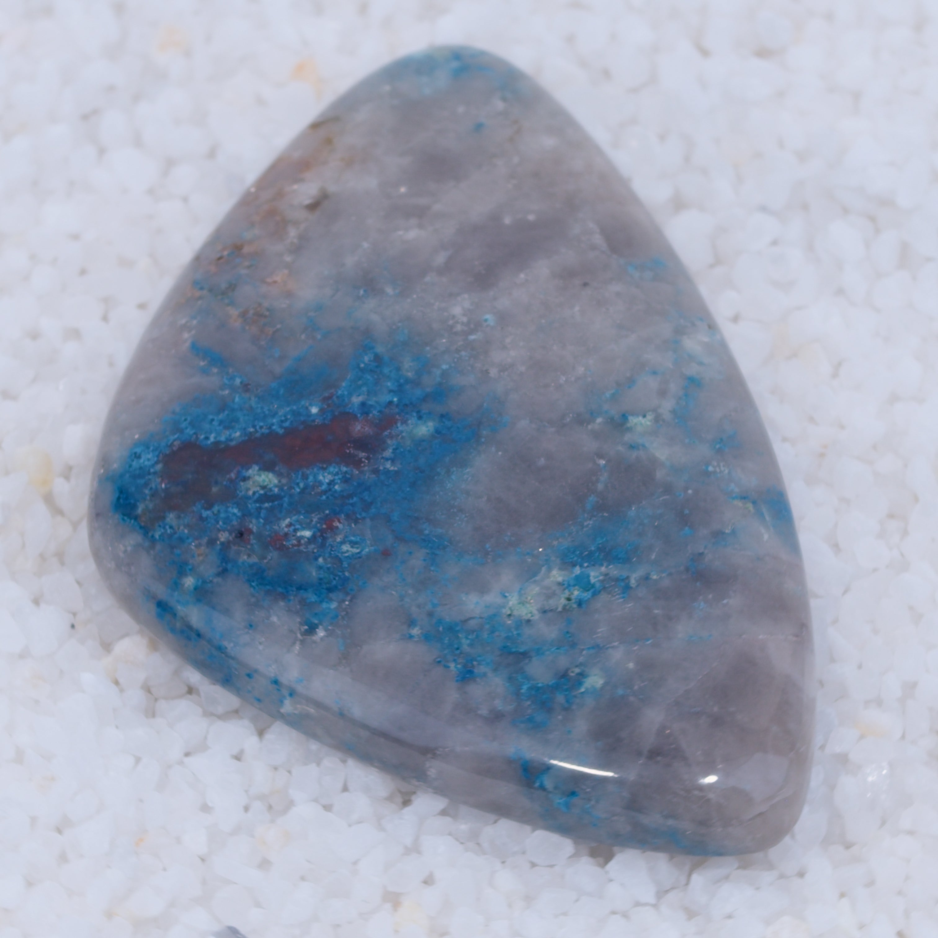 A polished, triangular Canyonite Sky LTD gemstone with a smooth surface features cloudy gray and vivid blue copper silicate patterns, creating a striking look against white pebbles. Irregular blue areas add to the gem’s unique appeal.