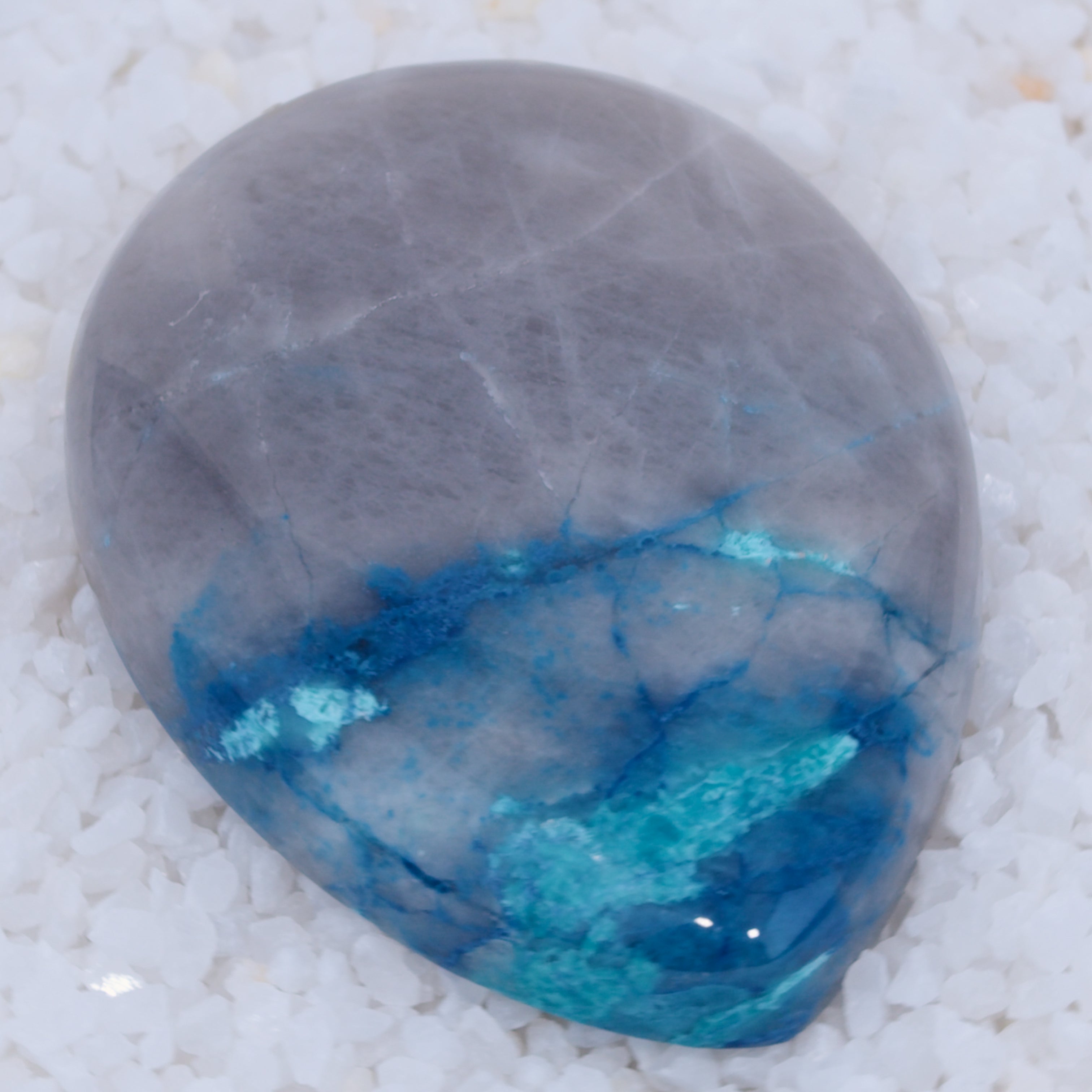 A smooth, polished teardrop-shaped Canyonite Sky LTD stone with a gradient from light gray to vibrant blue and rich in copper silicate minerals, resting on small white crystals—a striking gemstone for collectors.