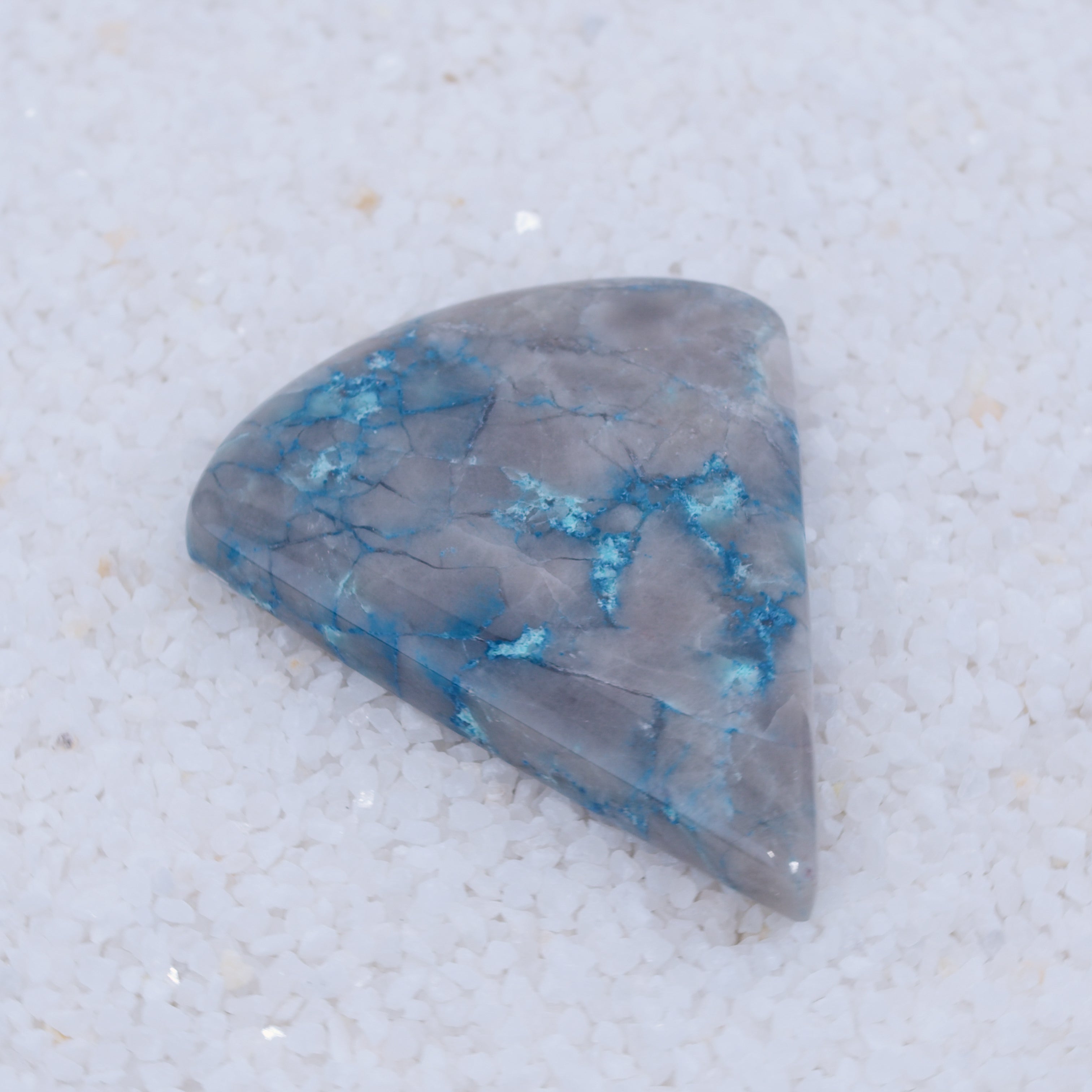 A polished, irregular gray stone from Canyonite Sky LTD, with bright blue veins of copper silicate, rests on white crystals—its vibrant patterns strikingly contrasted by the rough background.