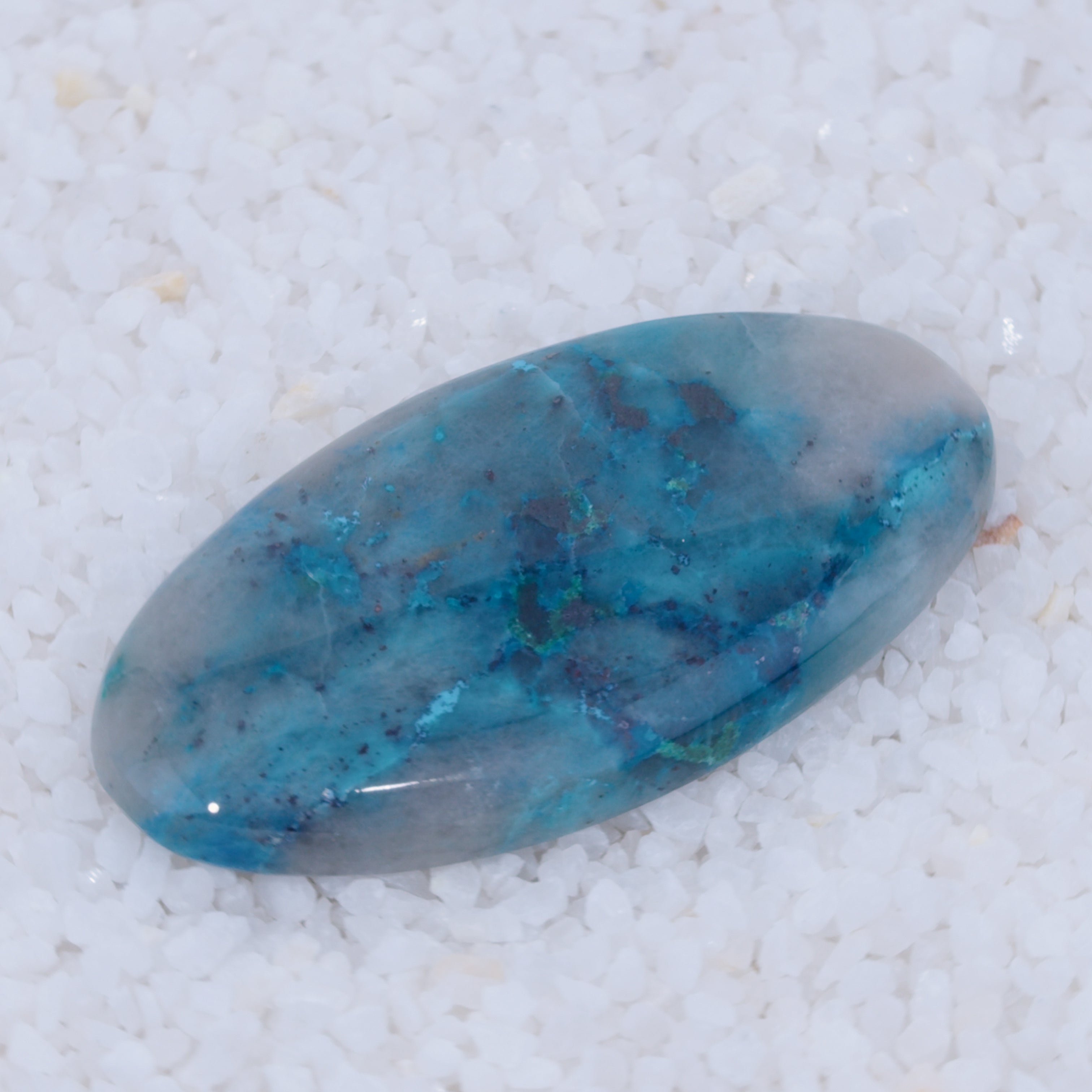 A polished, oval Canyonite Sky cabochon from Canyonite Sky LTD features marbled teal, blue, and gray swirls. Its glossy blue-green surface rests on smooth white pebbles—a distinctive collector’s gemstone.