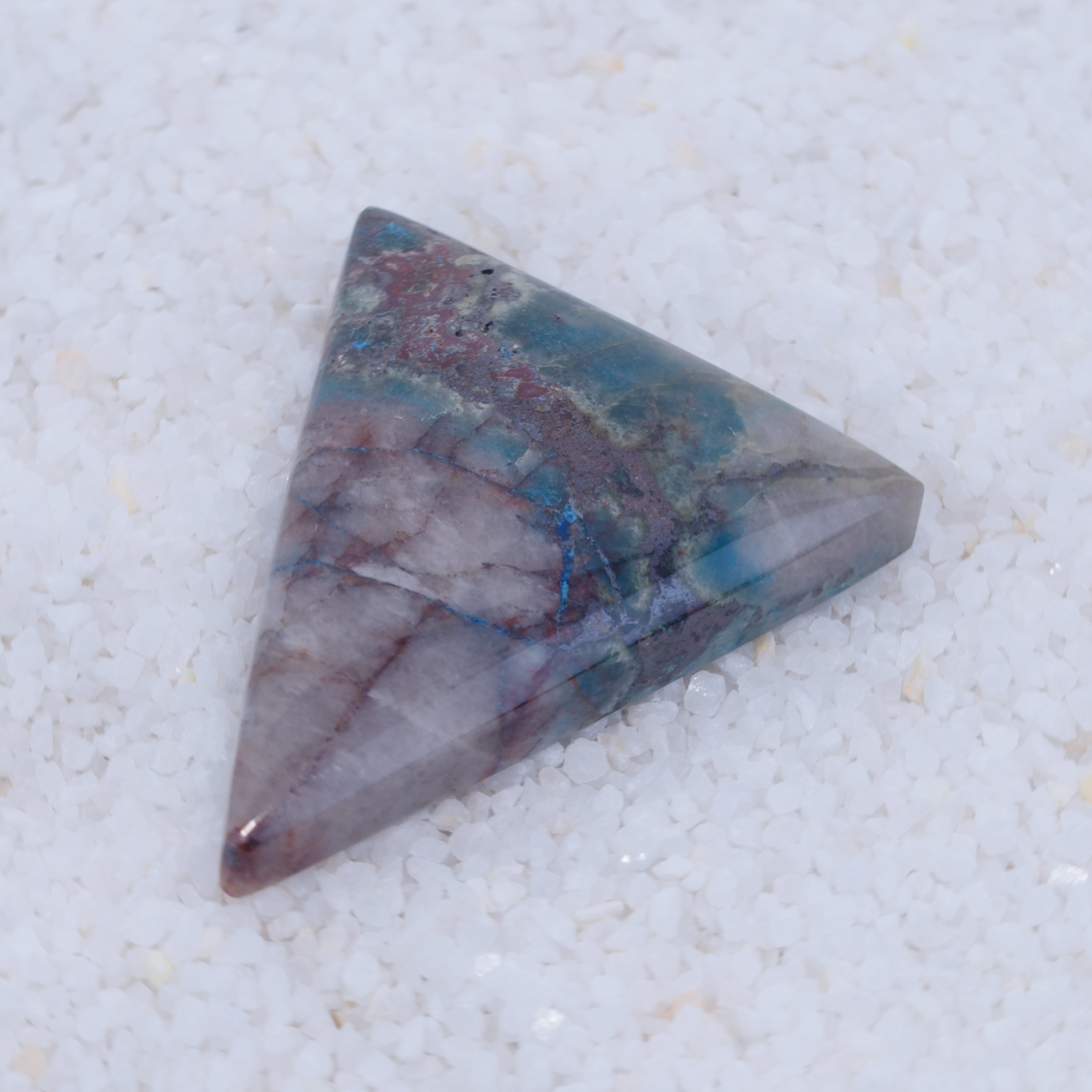 A polished triangular Canyonite Sky LTD cabochon displays blue, green, gray, and reddish-brown veining with varied patterns and natural inclusions on a smooth, glossy surface; a rare collector’s gemstone resting on white pebbles.
