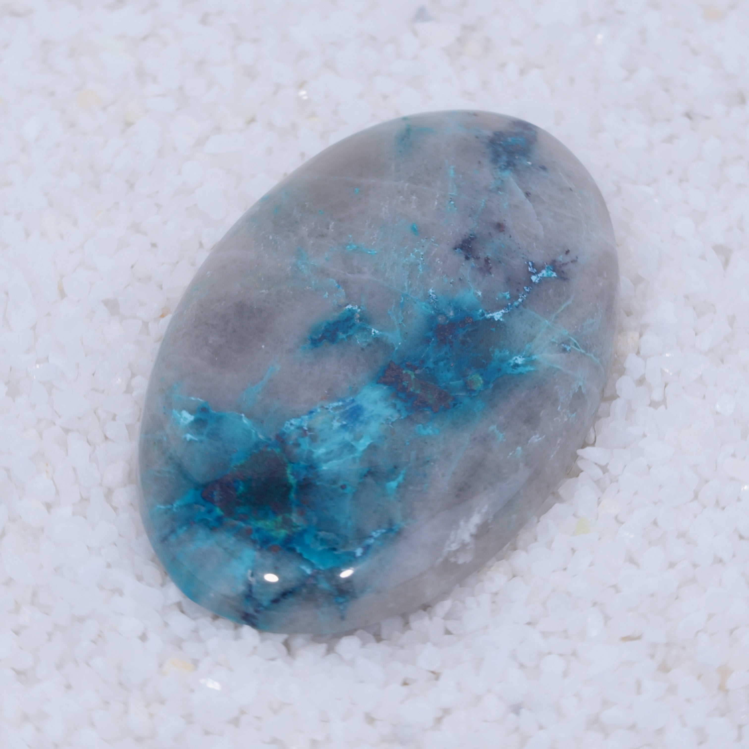 A smooth, oval Canyonite Sky LTD gemstone with a gray base and vibrant blue streaks lies on small white crystals—a striking contrast that makes this rare cabochon a true collector’s piece.