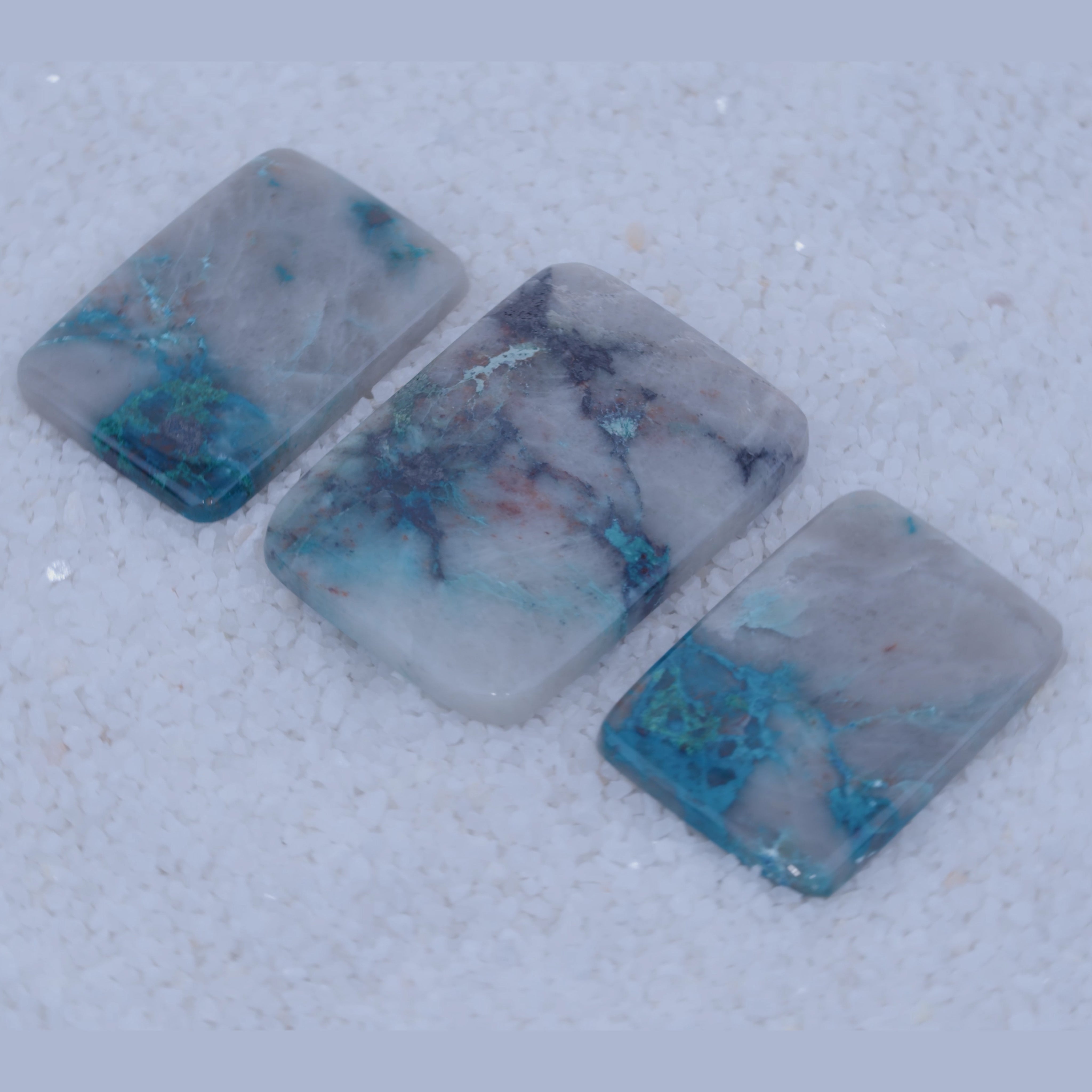 Three square stones with blue and green mineral inclusions on a textured gray surface
